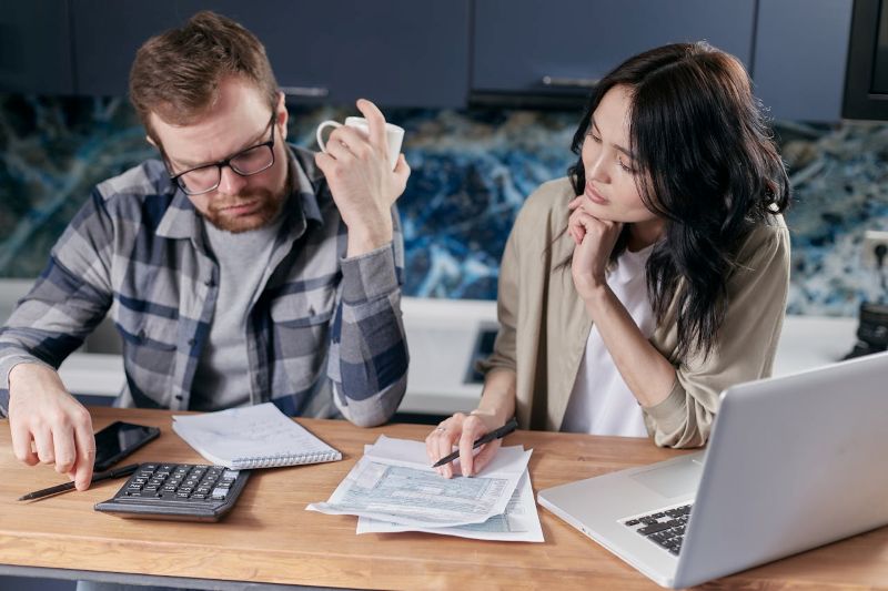 A man and a woman analyzing financial documents at a table, surrounded by a laptop and calculators, focused on business finance management.
