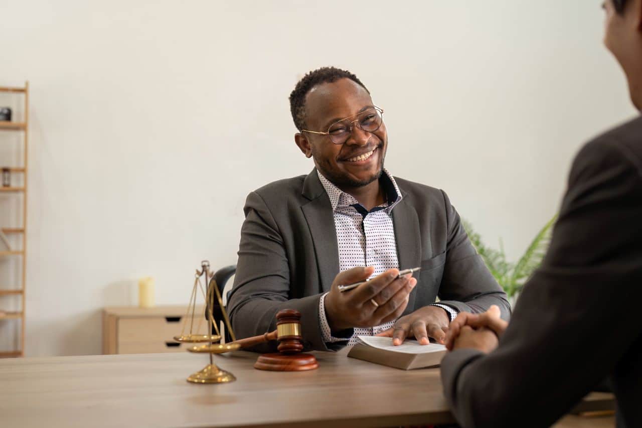 Smiling estate planning attorney sitting at a desk with legal scales and gavel, discussing documents with a client in a professional office setting.